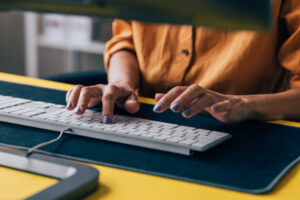 Close-up of a woman's hands typing on a white computer keyboard with a yellow desk.
